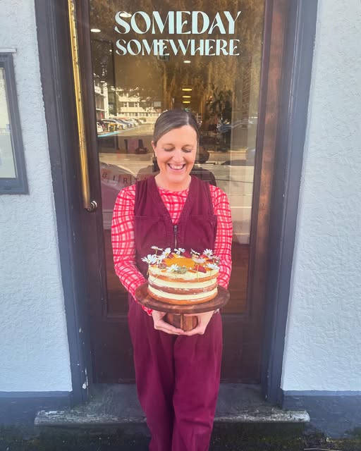 Kimberley Eagle smiling as she holds a layered celebration cake outside the café door.